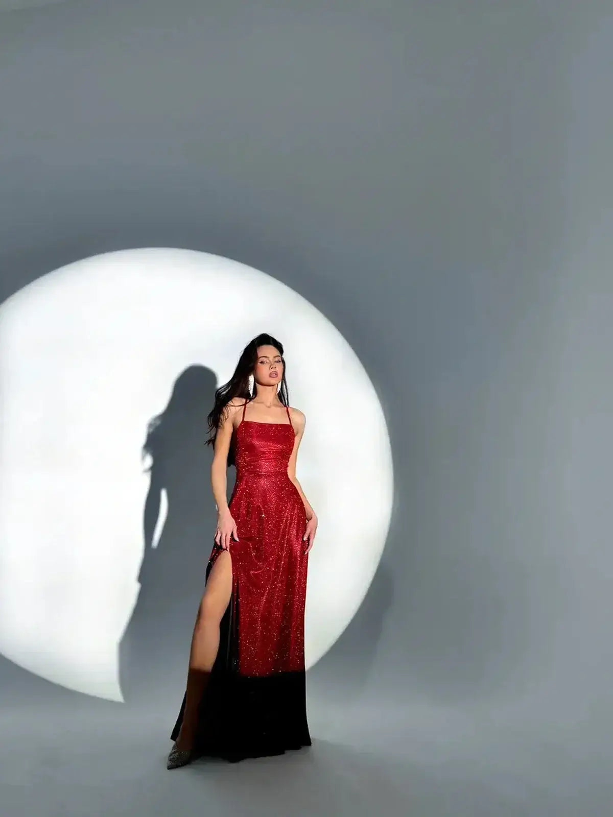 Model wearing a strappy red Maxi Dress Scarlett Shine with shimmer finish, showcasing a thigh-high slit against a dramatic backdrop.