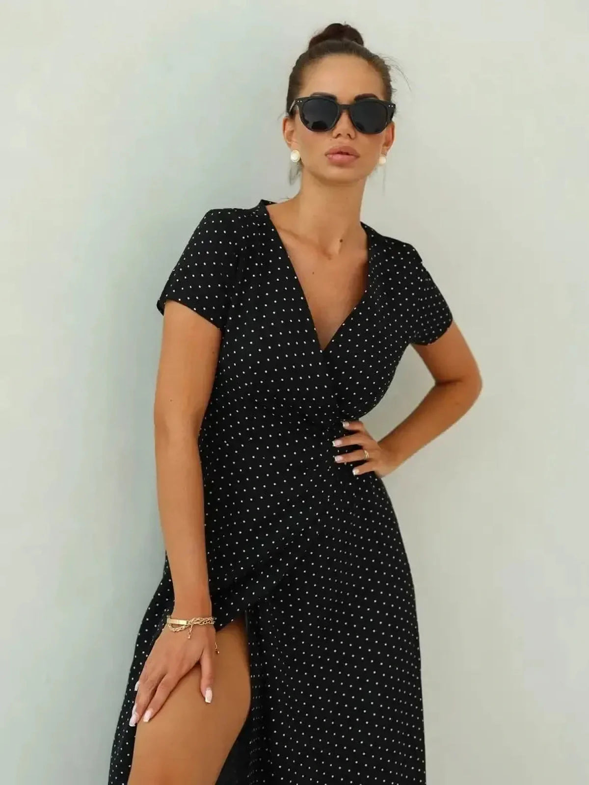 Stylish woman in a black polka dot maxi dress with wrap silhouette and thigh-high slit, wearing sunglasses.
