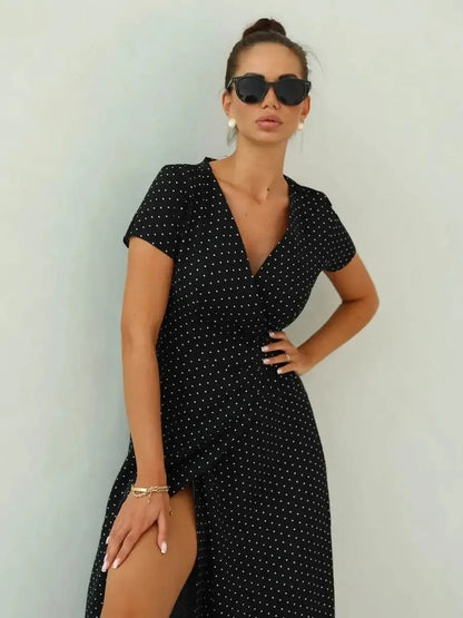 Stylish woman in a black polka dot maxi dress with wrap silhouette and thigh-high slit, wearing sunglasses.