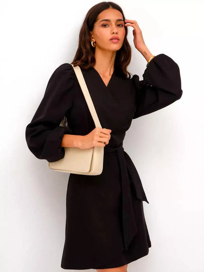 Model wearing a black mini dress with a V-neckline and flowing side ties, accessorized with a cream handbag.
