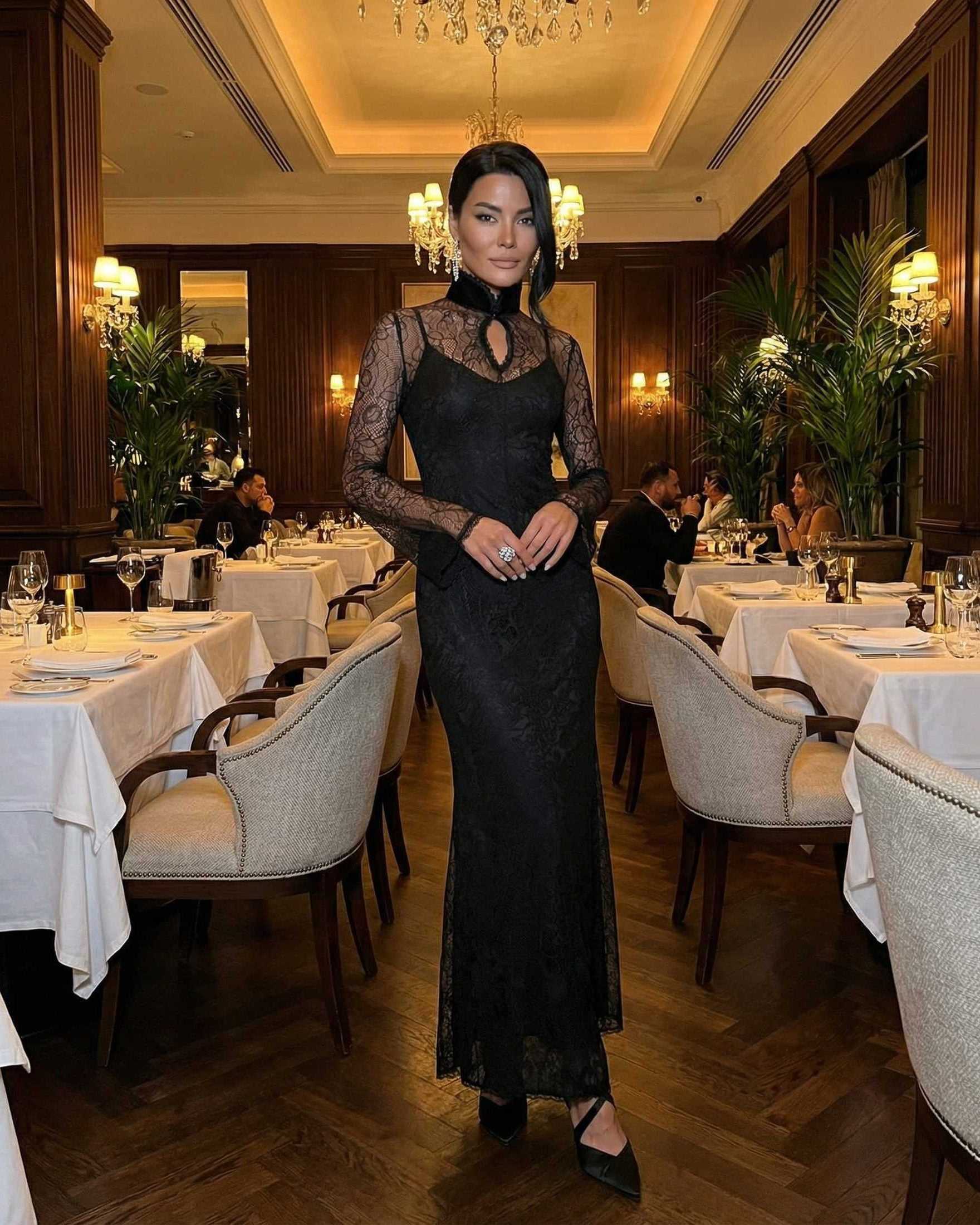 Woman wearing Noctavia Maxi Dress by Lauven, black lace long sleeve bodycon dress with stand collar in elegant restaurant.