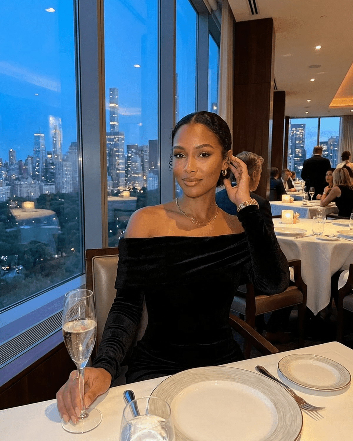 Elegant woman in Noctura Maxi Dress by Lauven, featuring off-shoulder design, seated at a table with city view.