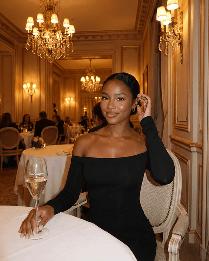 Elegant woman in Nocturne Maxi Dress by Lauven, off-shoulder style, seated in a luxurious dining setting.