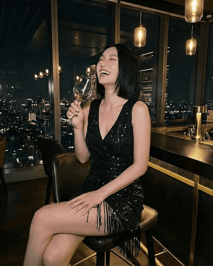 Lauven Noir Mini Dress worn by a woman laughing while holding a wine glass, featuring a black sequin design and fringe hem.