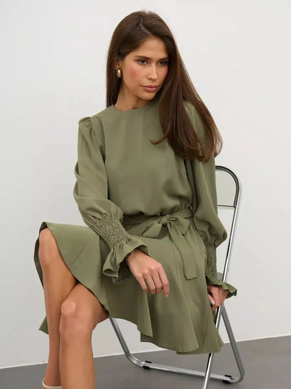 Model wearing the Olive Luxe Mini Dress in olive green, featuring long sleeves, ruffle details, and a waist-sash.