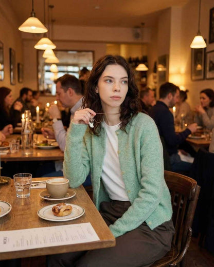 Sage Cardigan by Lauven worn by a woman in a cozy café setting, showcasing its stylish round neck and button front.