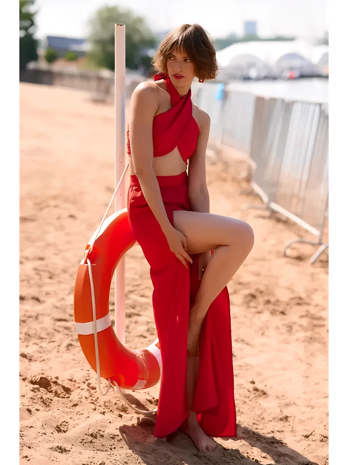 Model in a striking red summer two-piece set featuring a draped top and high-waisted slit skirt, posed by a lifebuoy.