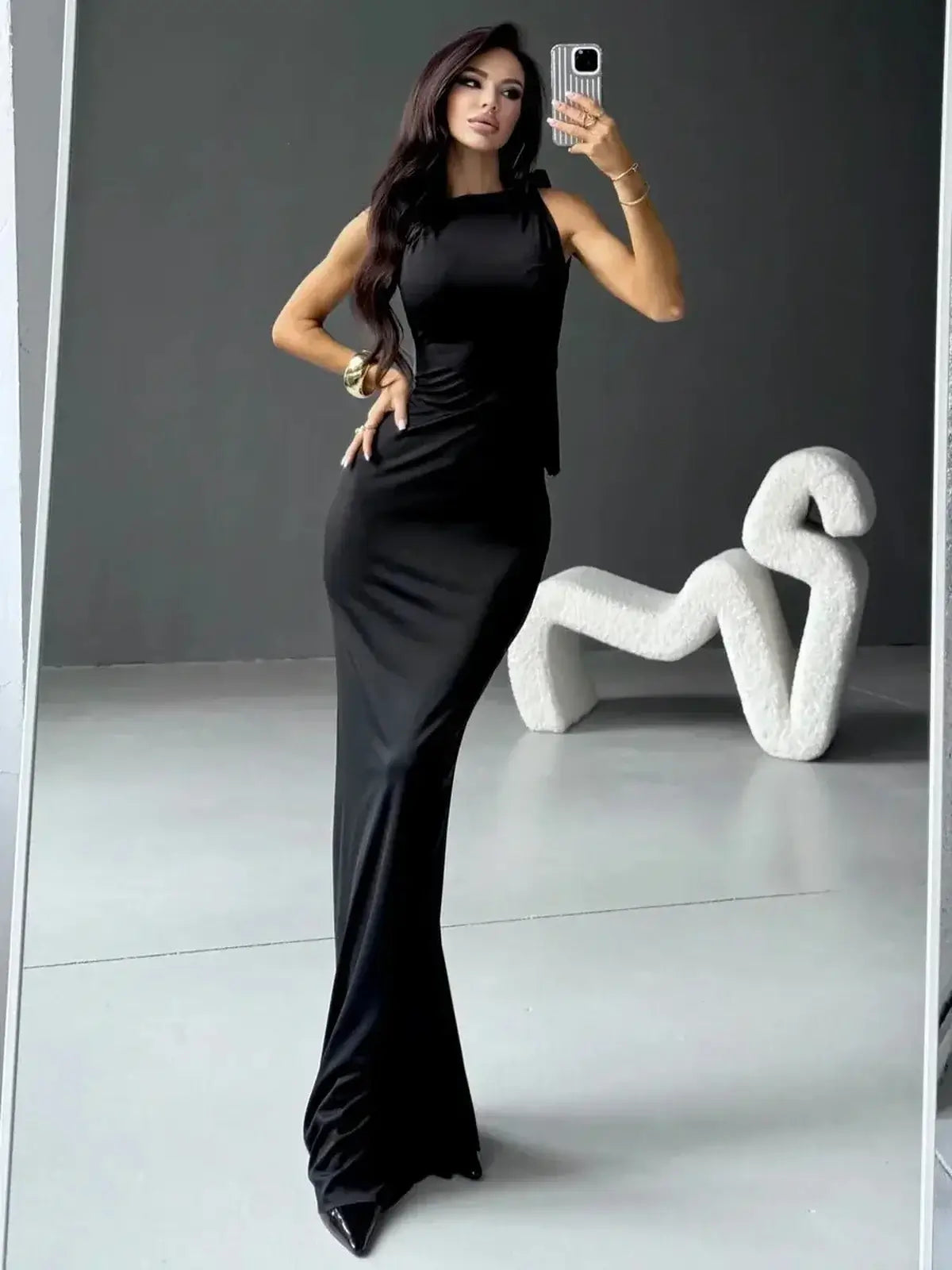 Elegant woman in Sculpted Noir Maxi Dress showcasing sleek silhouette and bow detail against a modern backdrop.