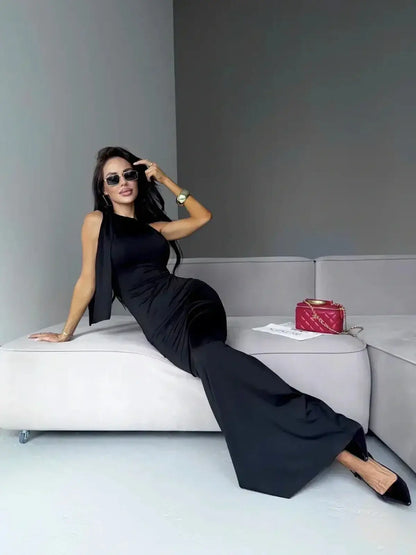 Model wearing a sleek black Sculpted Noir Maxi Dress, showcasing an elegant bow detail while relaxing on a stylish sofa.