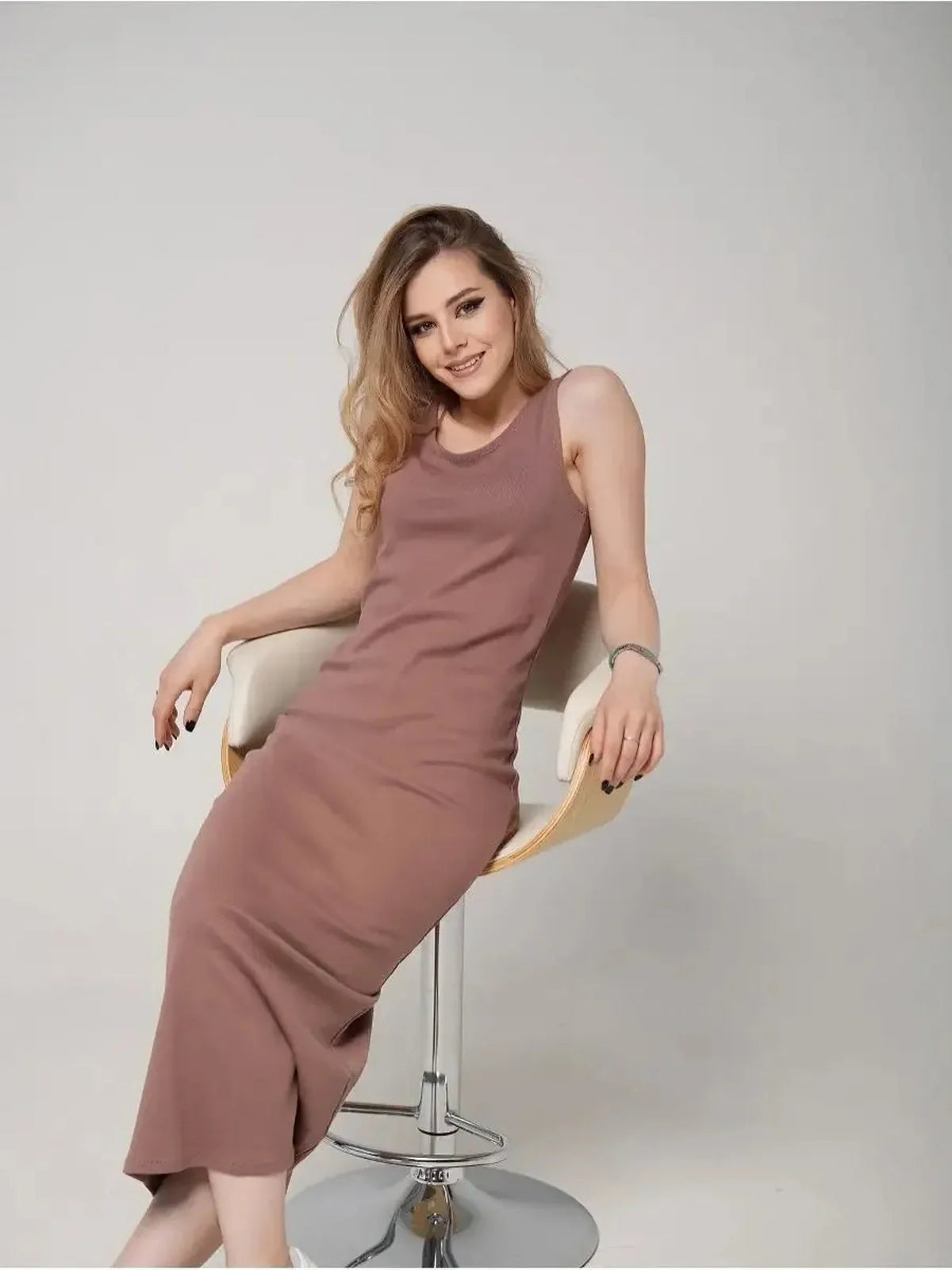 Model wearing a sleeveless Serene Flow Maxi Dress in a soft brown shade, seated elegantly on a stool.