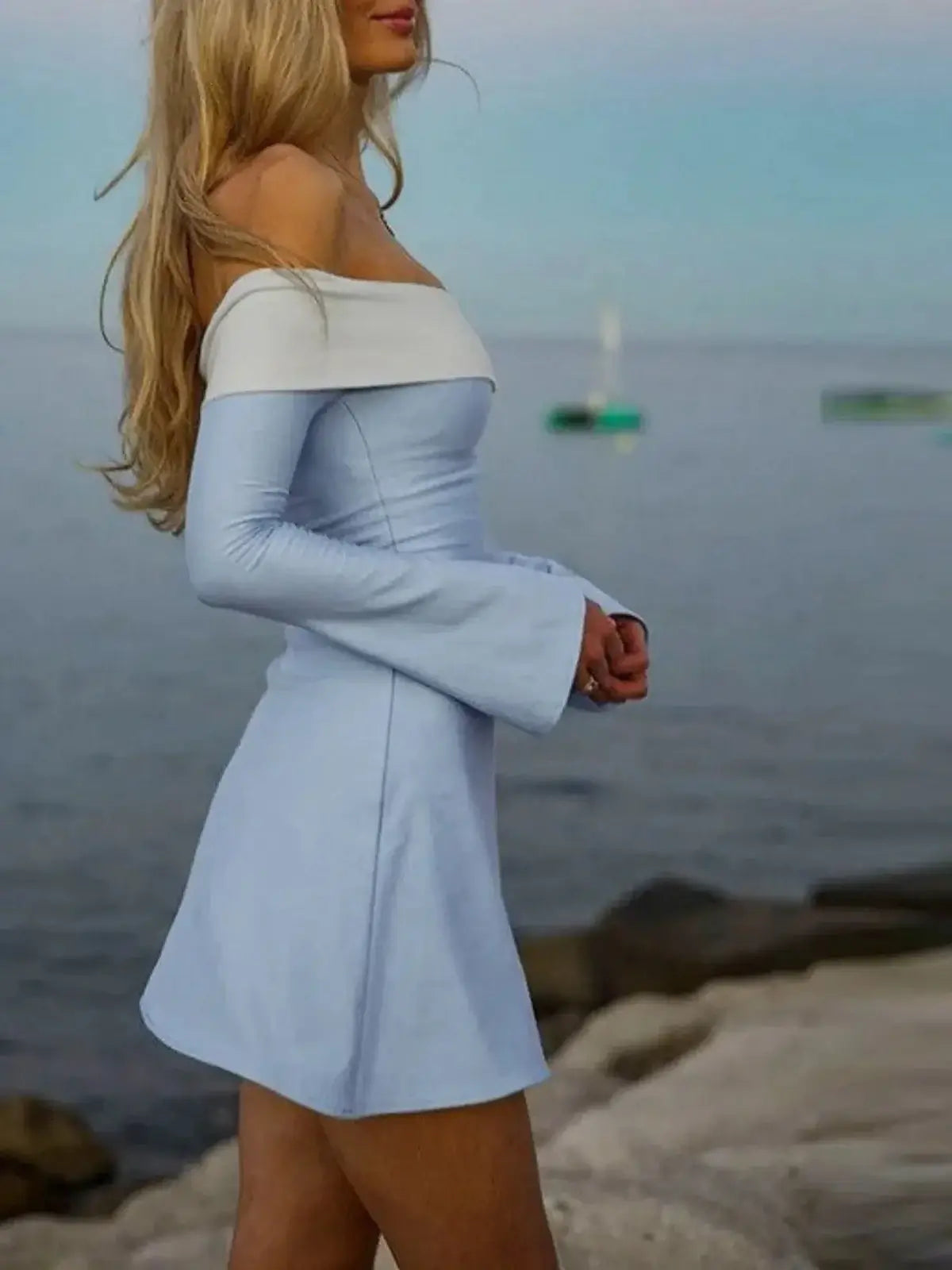 Woman in a sky blue off-the-shoulder mini dress by the water, showcasing a flattering silhouette and flared sleeves.
