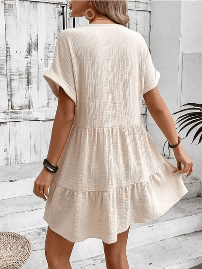 Back view of a woman in a beige breezy mini dress, showcasing its relaxed A-line silhouette and cuffed sleeves.