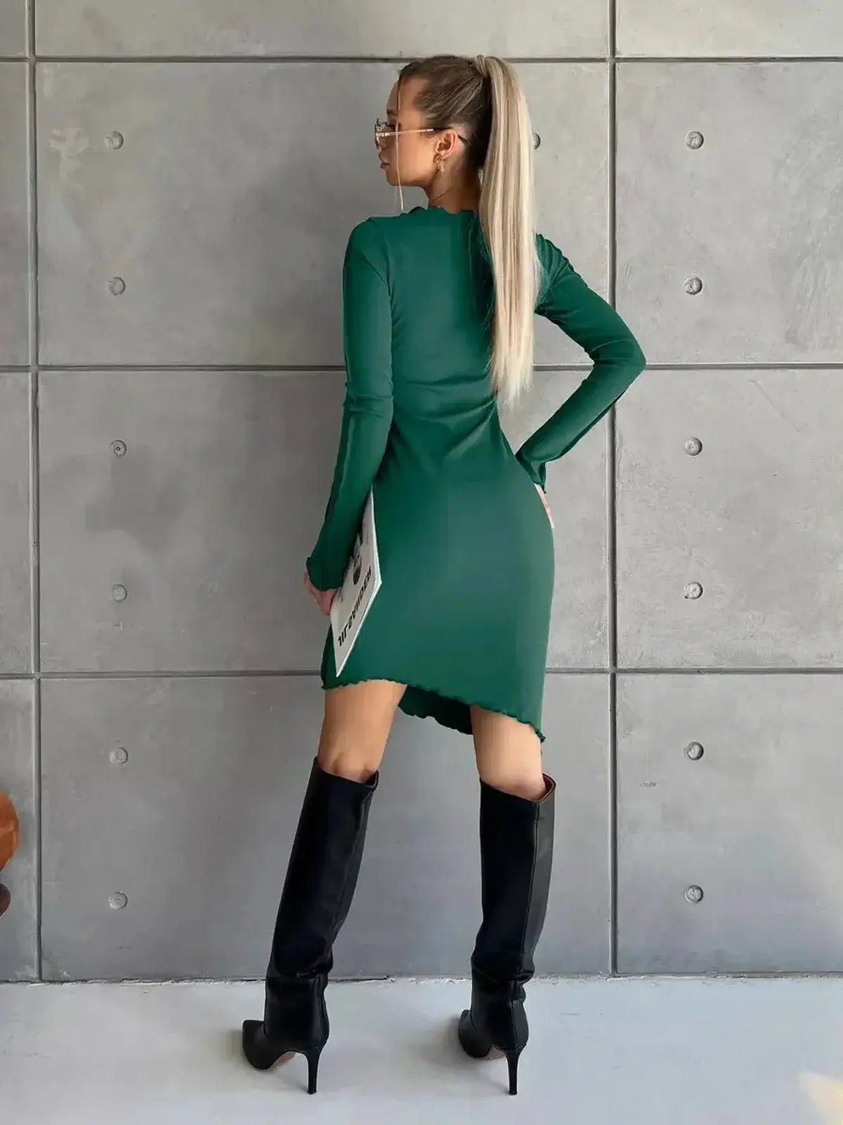 Fashion-forward model showcasing a long sleeve green mini dress with a stylish asymmetrical hem and sleek black knee-high boots.