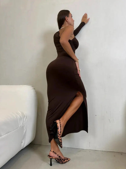 Model in a chocolate brown Velour Muse Maxi Dress with a high side slit and one-shoulder style, posing elegantly.