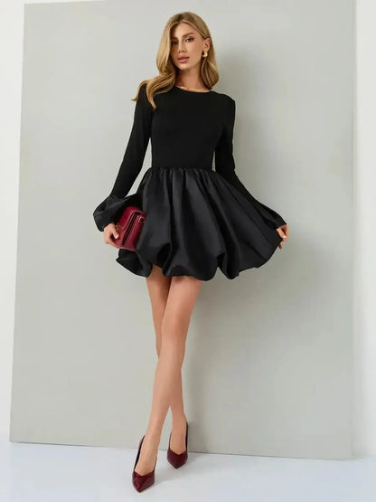 Model wearing Velvet Bloom black mini dress with satin balloon skirt and long sleeves, holding a red clutch.