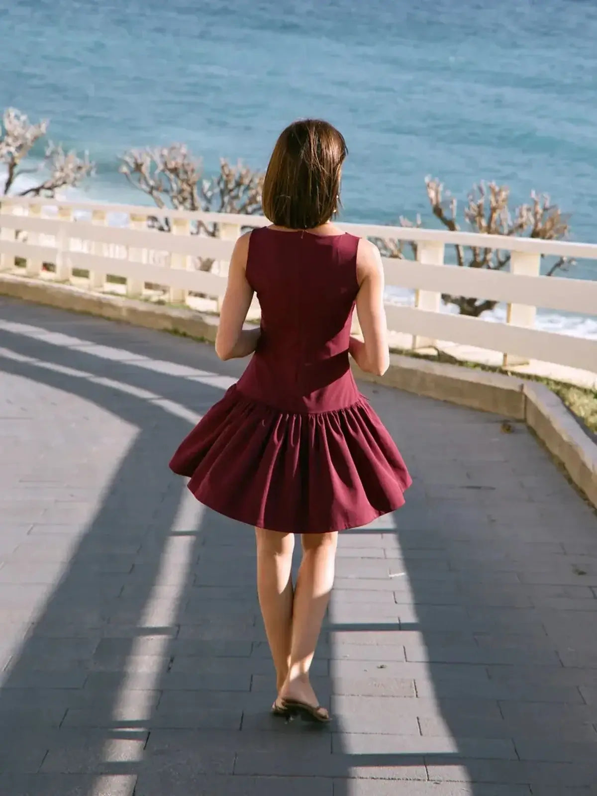 Model wearing a maroon Velvet Drop mini dress by the beach, showcasing a flowy pleated skirt and fitted bodice.