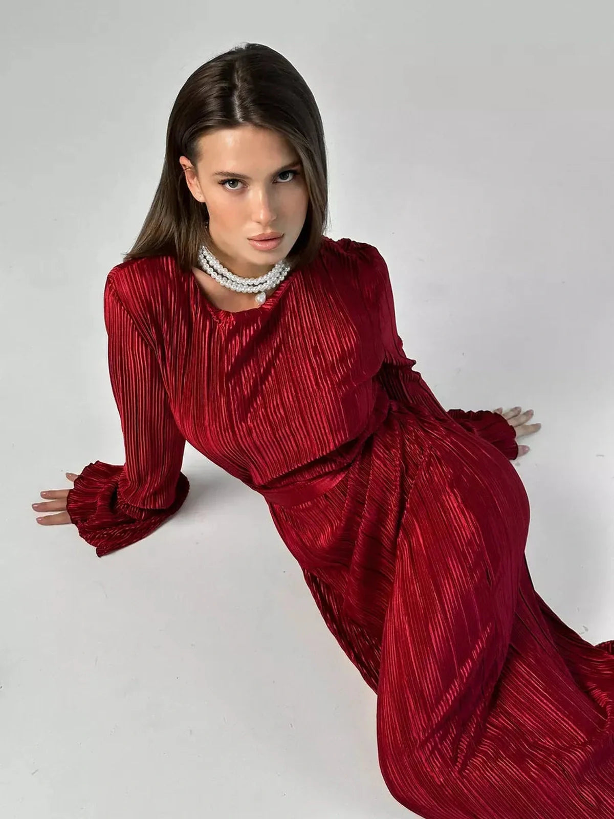 Elegant model in glossy Marsala red Velvet Flame Maxi Dress with pleats and flared sleeves, accessorized with a pearl necklace.